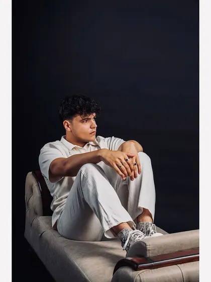 A full-body shot of a model in a relaxed, all-white outfit, seated against a dark background. The simple setup focuses all attention on the apparel and the model's contemplative pose.