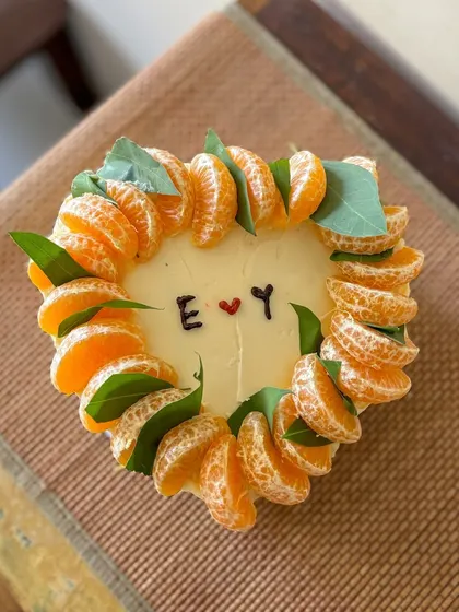 A rustic and minimal heart-shaped cake. It's decorated with a crown of fresh mandarin orange slices and leaves, with simple initials in the center for a personal touch.