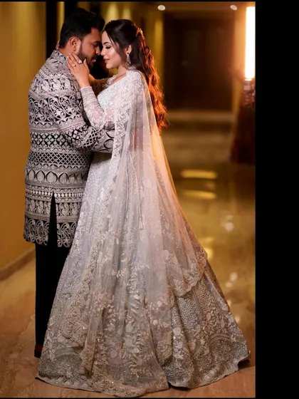 A full-length portrait in a beautifully lit corridor. The bride's flowing white lehenga and the groom's detailed sherwani create a stunning, elegant image of the couple.