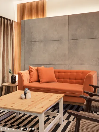This living area showcases a powerful contrast between a warm, vibrant orange sofa and a cool, industrial-style concrete panel wall. The striped rug adds another layer of bold pattern, creating a space that is both edgy and inviting.