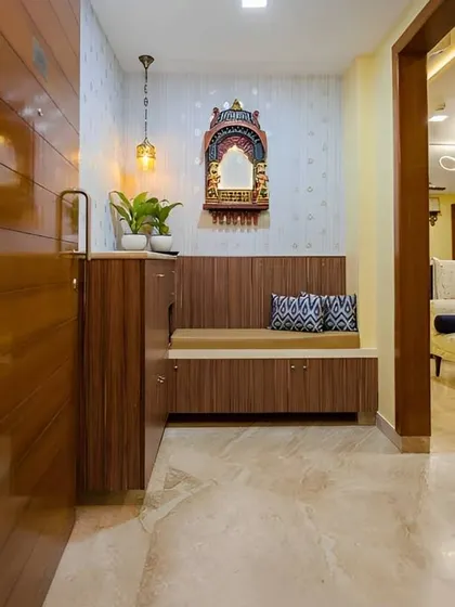 A traditional and cozy foyer with a wooden bench and shoe cabinet. A small, wall-mounted mandir adds a spiritual touch to the entrance.