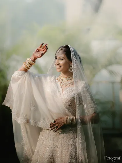 A happy bride in her all-white wedding lehenga, waving with a smile. The soft, diffused light gives the portrait an ethereal quality.
