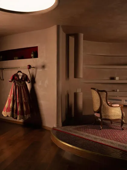 A bridal lehenga is displayed against a terracotta-hued wall, with a classic rattan chair and integrated shelving nearby. This composition balances the richness of the garment with the tactile, old-world nostalgia of the architectural finishes.