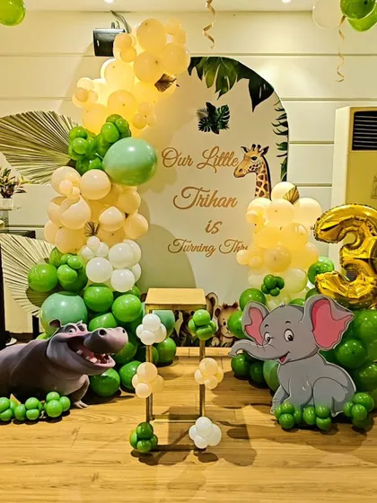 A cute jungle-themed decoration for a 3rd birthday. This setup includes a custom arched backdrop, elephant and hippo cutouts, and an organic balloon garland in green and yellow tones.