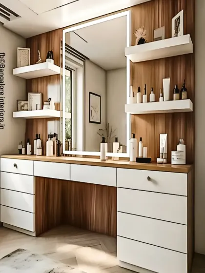 A modern dressing table with a large, backlit mirror providing perfect lighting. The unit offers extensive storage with multiple drawers in a clean white finish, contrasted with a wood-paneled wall and floating shelves for display.