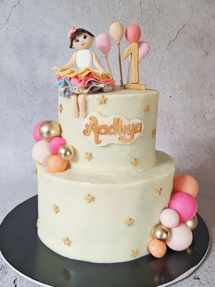 This two-tier cake is perfect for a first birthday, featuring a handcrafted fondant girl in a rainbow tutu. The cake is decorated with stars and colorful chocolate spheres for a fun, girly vibe.