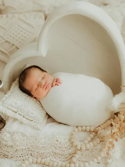 A newborn rests peacefully in a heart-shaped wooden bowl, swaddled in white. This setup uses natural textures and soft lighting for a pure and organic feel.