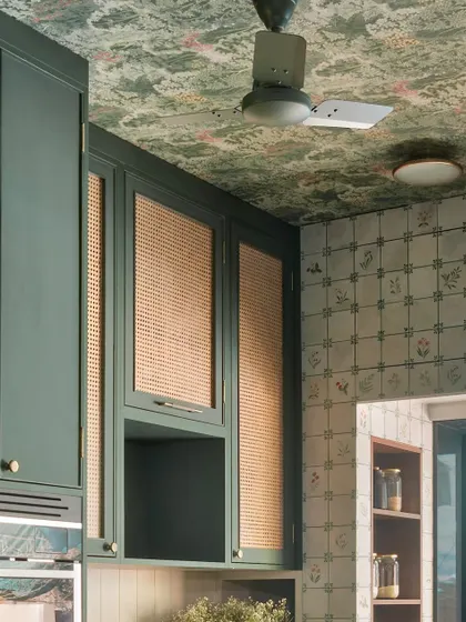 The ceiling wallpaper and patterned wall tiles create a rich tapestry of design in this kitchen. The wicker-fronted cabinets add another layer of texture, making the space feel deeply personal and curated.