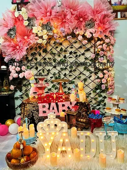A cute and cozy setup for a baby's birthday or naming ceremony. The decor includes a sequin backdrop, a floral and tulle arch, and light-up marquee letters spelling the baby's name.