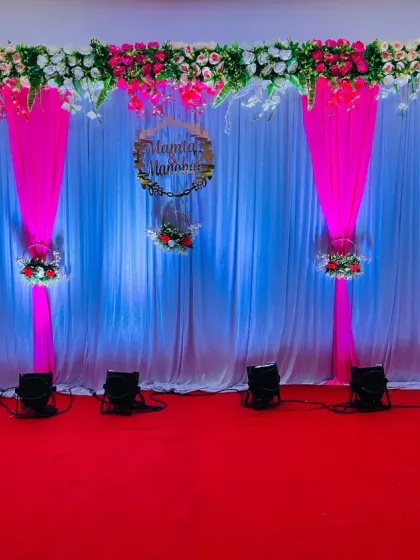 An elegant and simple stage decoration with a white curtain backdrop and tasteful pink drapes. A floral arrangement hangs from the top, and a custom wooden hoop with the couple's names adds a personal touch to this reception setup.