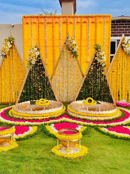 A truly unique Haldi seating arrangement. This setup features triangular backdrops made of marigold flowers and lush greenery, with intricate floral rangoli on the ground for a stunning visual effect.