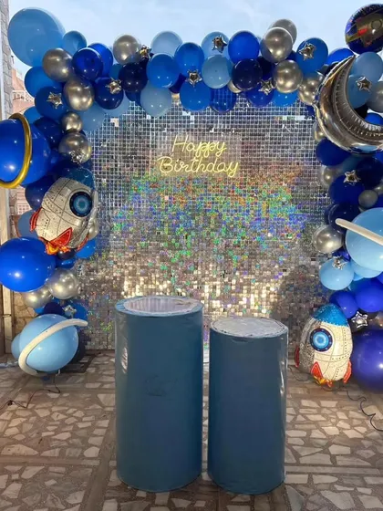An astronaut theme with a silver sequin backdrop creating a starry sky effect, framed by a blue and silver balloon arch with rocket and planet accents.
