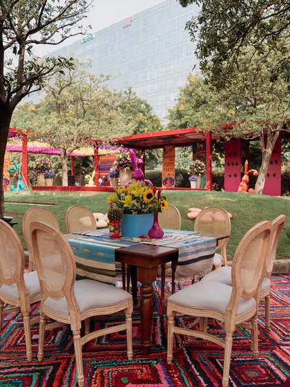 An outdoor dining area at the Mexican fiesta. The tables are set on a vibrant rug, and the surrounding trees and greenery create a beautiful garden party atmosphere.
