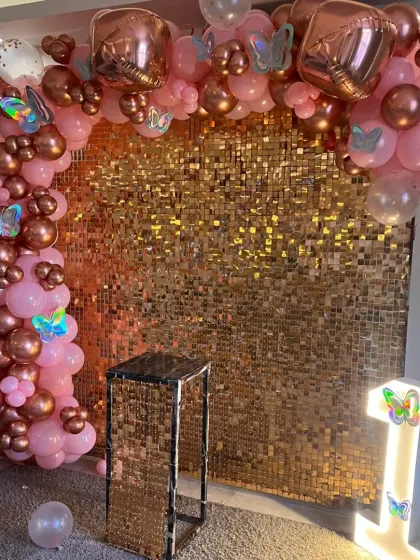 A beautiful first birthday setup with a rose gold sequin wall, a pink and chrome rose gold balloon arch, and a light-up number '1'. The butterfly accents add a whimsical touch.