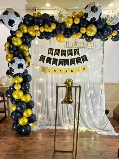 A football-themed party for a first birthday. The black and gold balloon arch is decorated with football balloons, set against a curtain with fairy lights.