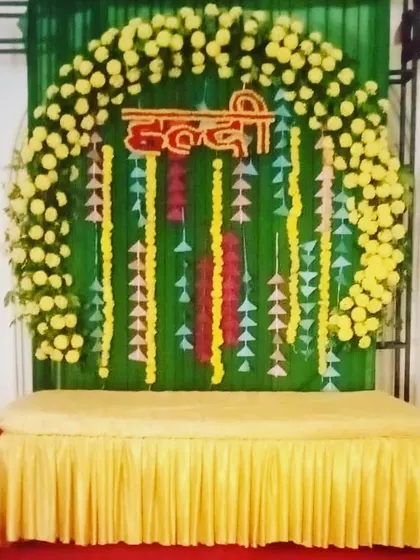 A Haldi backdrop with a circular arch of yellow flowers against a green curtain. The decor includes hanging paper craft for a simple, budget-friendly design.
