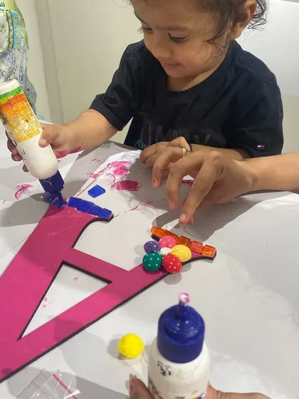 A toddler gets hands-on with glue and beads to decorate her initial. I ensure activities are safe and fun for even the littlest artists.