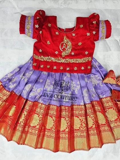 A custom pattu pavadai for a baby, featuring a personalized initial 'A' embroidered on the blouse. The red and lilac combination is rich and festive.