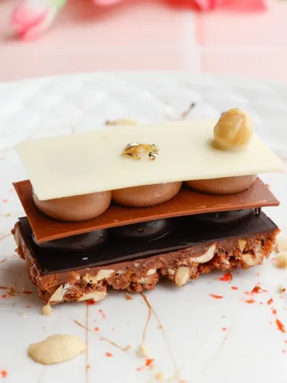 A detailed view of our Chocolate Mille-feuille. You can see the distinct layers of crisp pastry, smooth chocolate crémeux, and crunchy nuts, all topped with a delicate sheet of white chocolate.