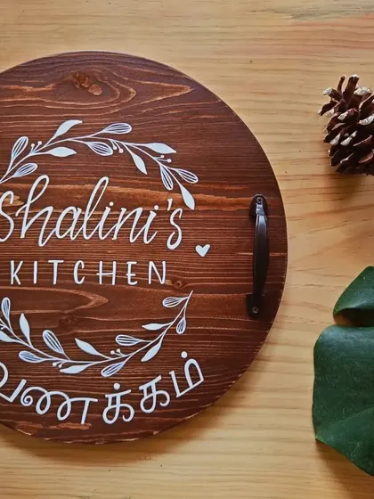 A beautiful, personalized kitchen board for "Shalini's Kitchen." The wreath design and "Vanakkam" greeting make it a warm and inviting piece for serving or display.