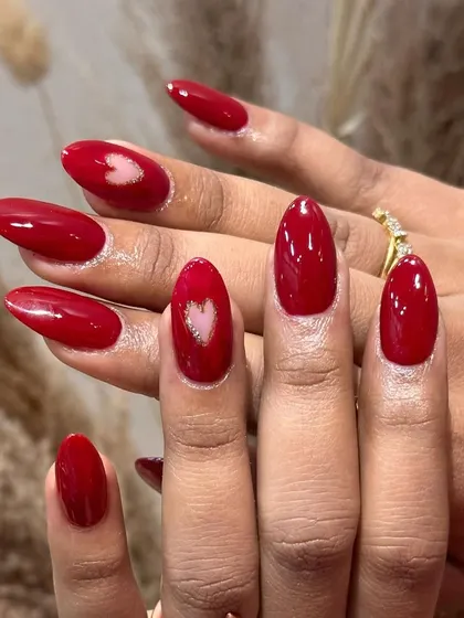 A full view of this romantic red manicure. The almond-shaped nails are painted in a vibrant red, with two accent nails featuring a beautiful, shimmering heart design.