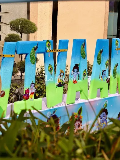 Large, custom-painted letters spelling 'VIVAAN' for a Krishna-themed party, each decorated with peacock feathers and images of Little Krishna.