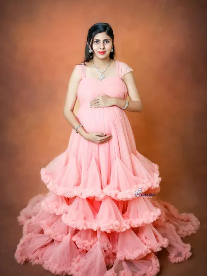 A full-length portrait of a mom-to-be in a beautiful tiered pink gown against a warm, neutral backdrop.
