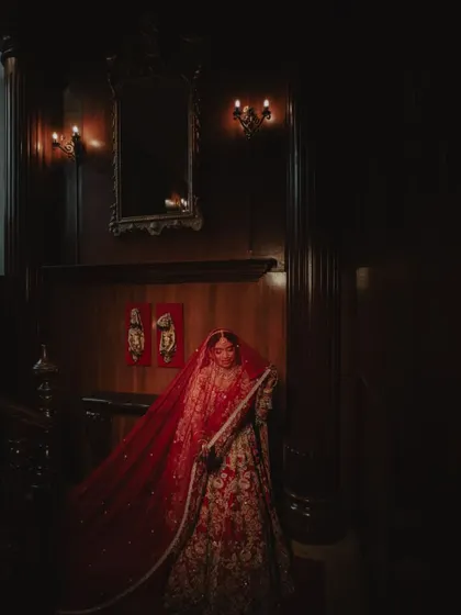 A dramatic portrait of the bride in a dark, wood-paneled room, her red lehenga illuminated by soft light.