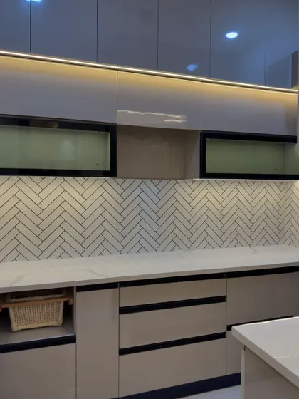 A simple and elegant kitchen with beige high-gloss cabinets and a white herringbone tile backsplash. The open basket storage is a practical touch.