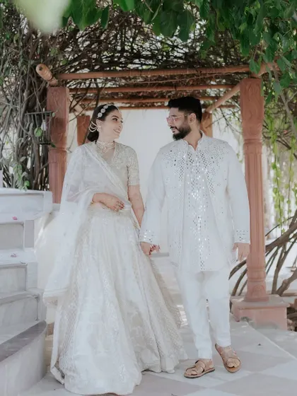 The couple walks hand-in-hand, sharing a look of love. The groom's mirror-work kurta adds a touch of traditional glamour to their coordinated white outfits.