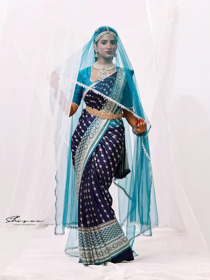 A powerful and direct pose, with the veil framing the model's face. This image highlights the striking combination of the dark saree and the light, airy veil.