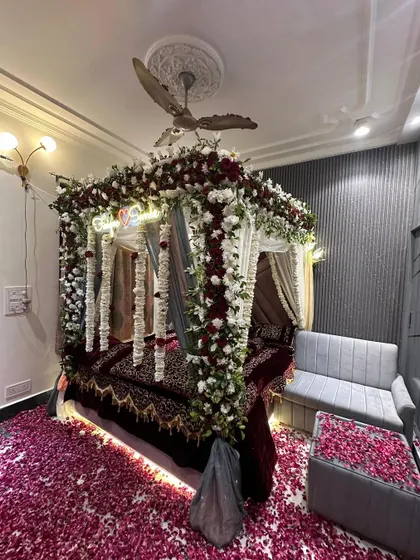 A luxurious 'Welcome Bride' setup with a four-poster bed heavily draped in red and white flowers. The floor is completely covered in red rose petals.