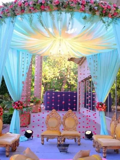 A dreamy mandap setup with pastel blue drapery and golden chairs. The canopy is adorned with pink and white flowers, and the floor is scattered with petals, perfect for a fairytale wedding.