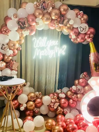 Another beautiful proposal decor idea. This one uses a circular balloon arch in rose gold and white, with butterfly accents, a champagne bottle prop, and a 'Will you Marry Me?' sign.