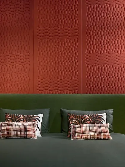 The 'Wave Flanelle' 3D wallcovering in a bold terracotta red, used as a headboard. The pure wool material and quilted undulations create a tactile, visually appealing surface with excellent acoustic properties.