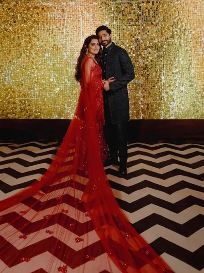 The couple poses against a black and white chevron floor and a gold sequin wall, a perfect example of our bold and glamorous Sangeet decor. The bride's dramatic red train adds to the stunning visual.