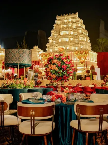 Guest dining tables with modern chairs and jewel-toned velvet tablecloths, set against the backdrop of the grand temple mandap. This view shows the thoughtful planning of guest experience and comfort.