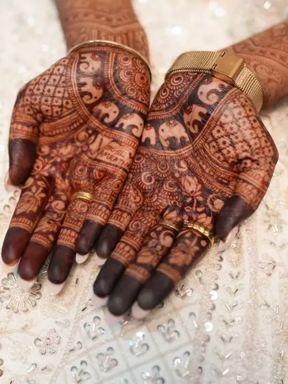 A close-up of a bride's hands, featuring detailed elephant motifs and geometric patterns. The dark stain looks incredible against her white wedding attire.