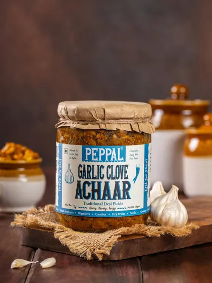 A product shot of Peppal's Garlic Clove Achaar, styled with fresh garlic and rustic pottery. The composition reinforces the traditional, flavorful nature of the pickle.