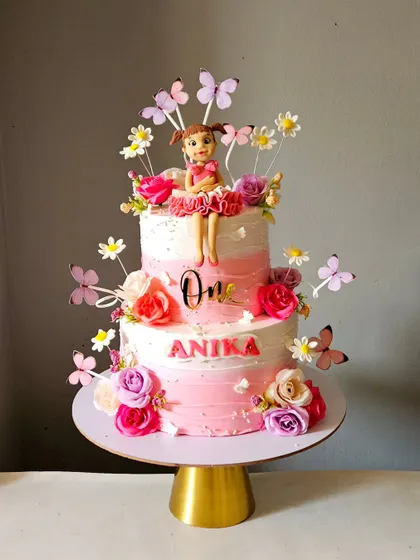 All things beautiful and dreamy for a first birthday. This two-tier cake features an ombre pink effect, a handmade doll topper, and is decorated with flowers and butterflies.