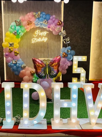 A butterfly-themed birthday party with large light-up letters spelling the name "RIDHVI". The setup includes a pastel-colored balloon ring and a light-up number "5".