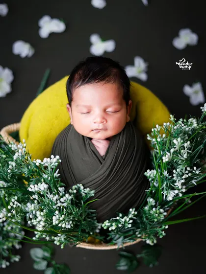 A baby swaddled in a dark wrap, nestled in a basket surrounded by delicate white flowers.