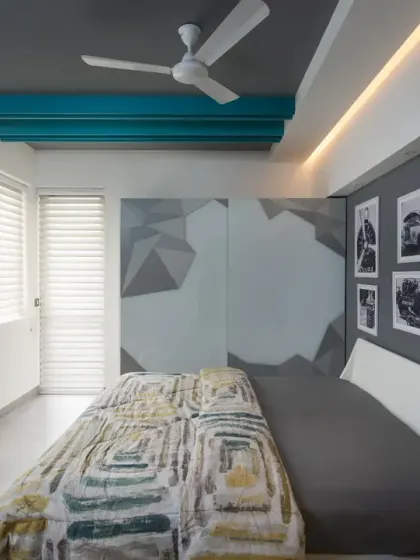 The guest bedroom in the Grey Apartment exudes a modern, clean aesthetic. Teal ceiling beams and a geometric accent wall introduce a bold visual contrast, while black-and-white photographs lend an artistic touch.