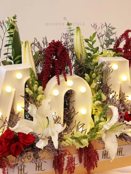 A close-up of the "MOM" floral hamper, showing the detail of the light-up letters and the lush arrangement of lilies and roses.