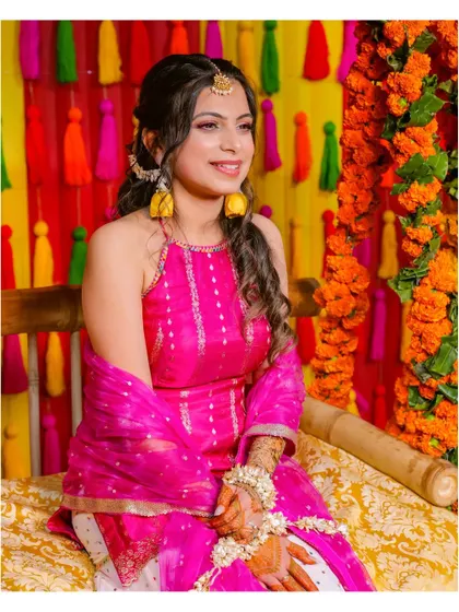 A portrait of the bride at her Mehendi ceremony, seated against a colorful backdrop of tassels. Her pink outfit and floral jewelry complete the festive look.