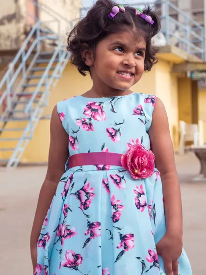 A beautiful sky blue dress with a vibrant pink floral print. The contrasting pink sash with a flower detail makes the colors pop, creating a cheerful and stylish look.