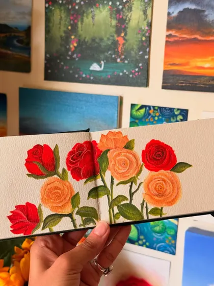A detailed shot of the rose painting in my sketchbook. You can see the texture of the paper and the fine brushstrokes I use to define each petal, with some of my other landscape paintings in the background.