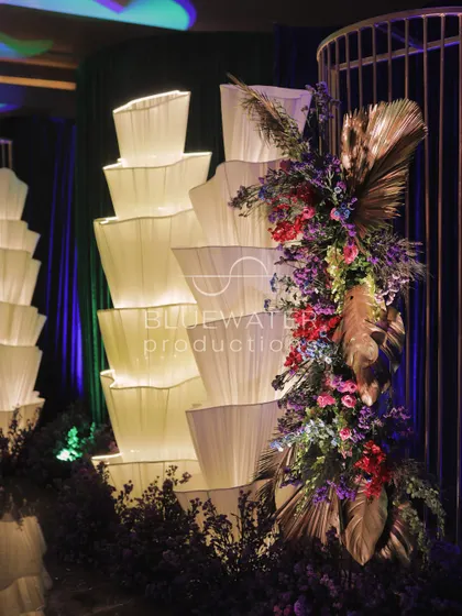 An artful floral installation combining oversized metallic leaves with vibrant purple and red blooms, placed next to a tower of soft, glowing fabric lights.
