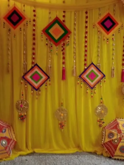 A traditional Haldi backdrop using bright yellow fabric drapes. The decor is accented with colorful hanging threadwork panels and decorative umbrellas, giving it a vibrant and festive feel.
