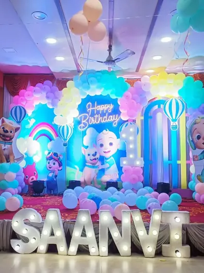 A colorful Cocomelon themed first birthday party. The stage is filled with characters from the show, a rainbow, and pastel balloons, along with large marquee letters for the name "SAANVI."